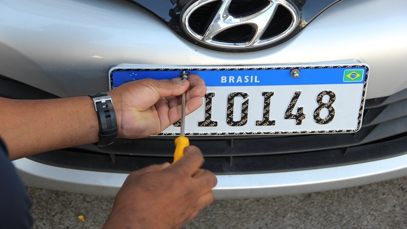 Foto da frente de um automóvel onde está sendo parafusada a placa de identificação, no padrão Mercosul (chapa branca com tarja azul no alto e números e letras pretas)