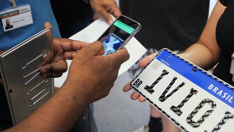 Mãos seguram placa veicular do novo modelo padrão Mercosul e outras mãos apontam o visor do celular para o QR Code da placa