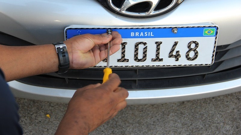 Foto de uma placa veicular sendo fixada na dianteira de um automóvel. Aparece apenas as mãos da pessoa que está aparafusando o dispositivo