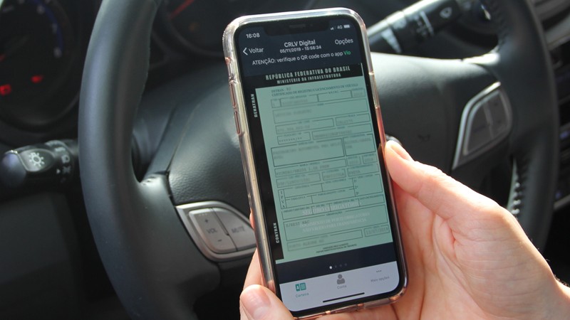 Foto de um condutor sentado ao volante, segurando o celular que na tela mostra o CRLV Digital