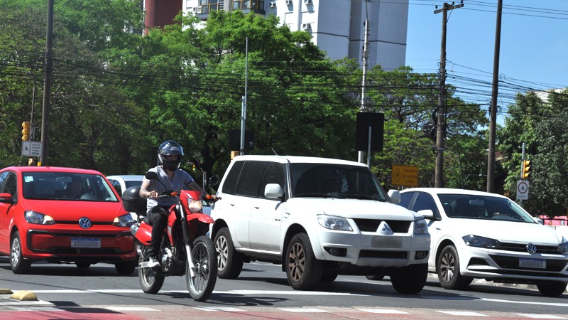 Foto diurna de uma situação de trânsito urbano. Mostra carros e motos parados antes da faixa de retenção, em um semáforo