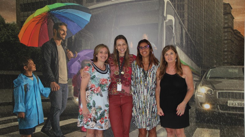 Alisandra Battistel com as servidoras do DetranRS Tatiane Stein, Adriana Reston e a diretora Diza Gonzaga. No fundo, a imagem de um painel que mostra a foto de um homem atravessando a rua na faixa de pedestres com duas crianças, enm um dia chuvoso.