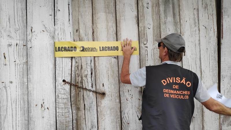 Servidor com colete da Divisão de Desmanches de Veículos do DetranRS cola uma faixa de lacrado na parede do estabelecimento clandestino