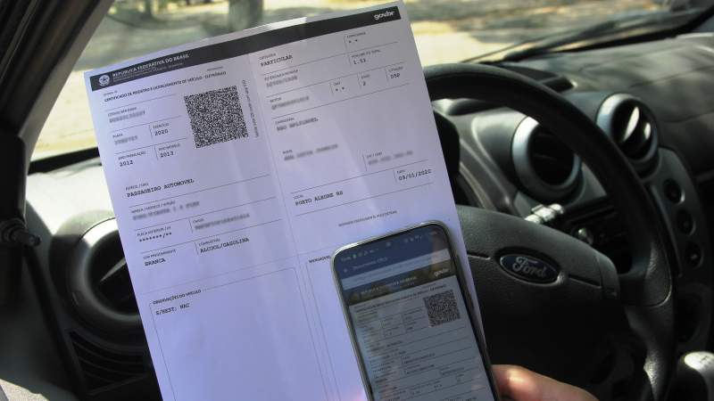 Foto que mostra um condutor sentado ao volante, segurando na mão as duas versões do CRLV-e, a versão imprimível, e também na forma digital