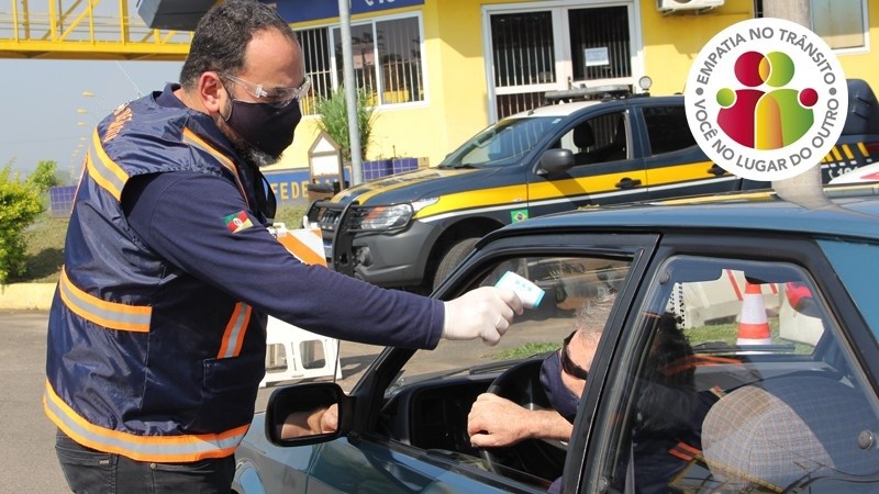 Agente de fiscalização do DetranRS mede a temperatura de um condutor abordado na Operação Empatia no Trânsito. O Veículo está estacionado em frente ao posto policial da PRF, à luz do dia. Condutor sentado no banco do motorista e o agente de pé ao lado.