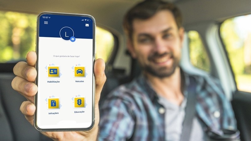 Homem jovem sentado em um carro mostra a tela do celular onde está aberto o aplicativo Carteira Digital de Trânsito (CDT), onde condutores e proprietários de veículos podem aderir ao Sistema de Notificação Eletrônica (SNE)