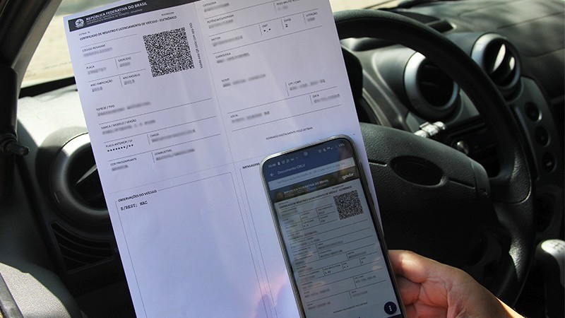 Na frente de um volante, motorista segura celular com o documento digital e papel com o documento impresso