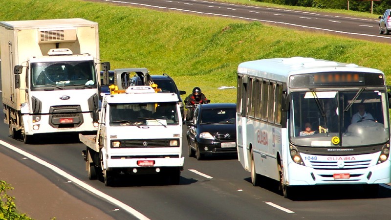 Foto de veículos pesados circulando junto com veículos leves em rodovia de pista dupla, durante o dia.