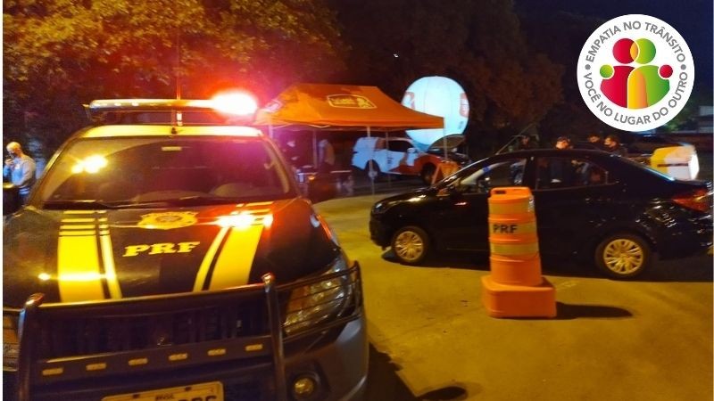 Foto noturna de blitz da Balada Segura, que mostra viatura da PRF parada com sinalização por giroflex ligada. Ao fundo a tenda da Balada Segura. Veículos estão parados na blitz enquanto seus condutores conversam com as autoridades