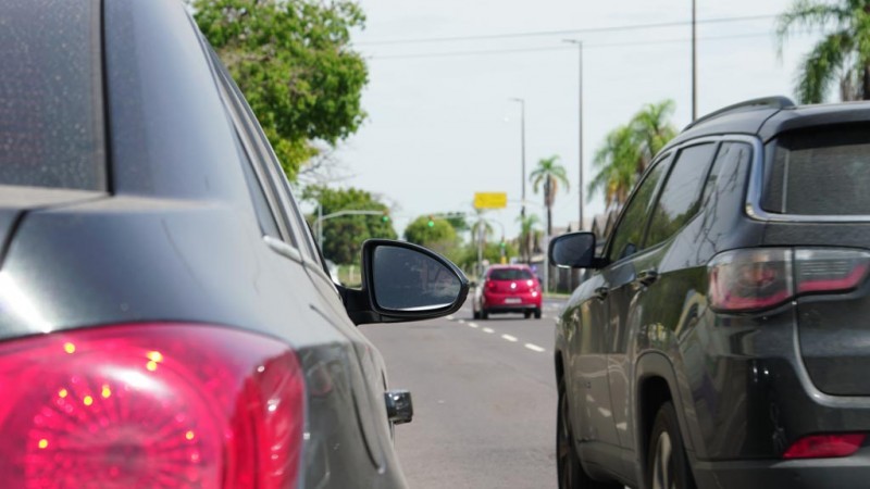 Imagem de dois veículos lado a lado, com a sinaleira traseira direita do veículo à direita em primeiro plano e a sinaleira traseira esquerda do veículo à esquerda um pouco à frente. Entre os dois carros, se vê outro veículo mais adiante na pista.