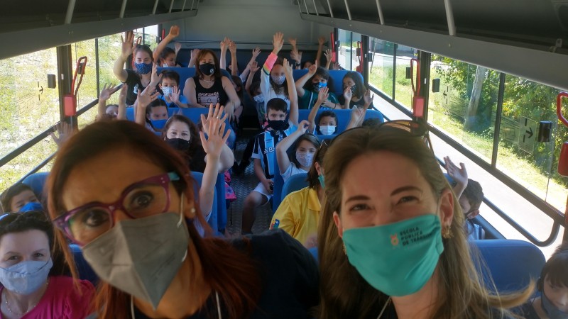 Duas mulheres de máscara sorriem para a camera dentro de um ônibus. Ao fundo, sentadas, crianças sorriem com os braços levantados