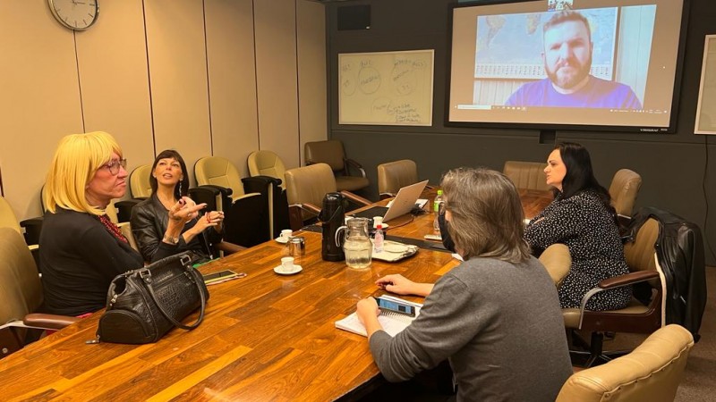 Três mulheres e um homem conversam em uma mesa de reunião e um homem participa online em um telão