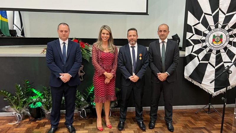 Foto colorida horizontal mostra três homens de terno e uma mulher loira de vestido vermelho ao lado de um brasão da Polícia Civil