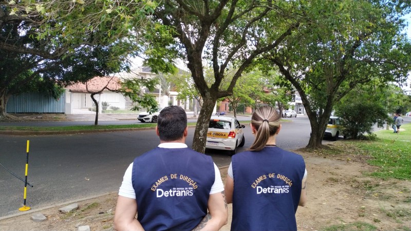 Foto horizontal colorida mostra homem e mulher de costas vestindo coletes de examinador do Detran e observando à distância um carro de autoescola fazendo a baliza em uma rua arborizada