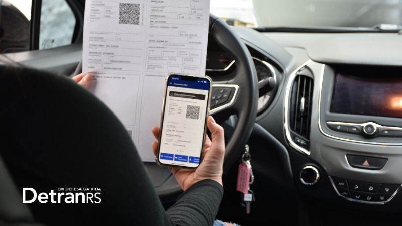 Foto horizontal colorida mostra motorista ao volante de um carro segurando um celular com o CRLVe na tela e uma folha A4 com o documento impresso