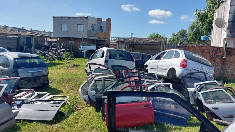 Foto diurna que mostra carcaças e peças de veículos dispostas em um terreno com gramado e cercado por muro de tijolos