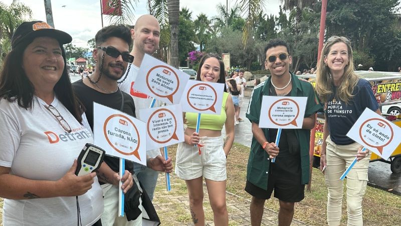 Foto diurna que mostra os servidores do DetranRS e alguns jovens abordados na ação educativa, em pé, posicionados lado a lado e segurando plaquinhas com mensagens educativas de trânsito