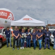 Foto diurna da equipe do DetranRS que atua na Balada Segura em pé, lado a lado, na frente da tenda, entre duas viaturas