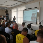 Diretor-geral adjunto Rafael Mennet ao microfone, fazendo uma explanação sobre o trabalho da Balada Segura na operação RS Verão Total. O público, sentado, observa o que aparece no telão.