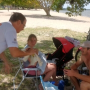 Família sentada à sombra de uma árvore na beira da prainha da Lagoa dos Patos recebe orientações do servidor do DetranRS