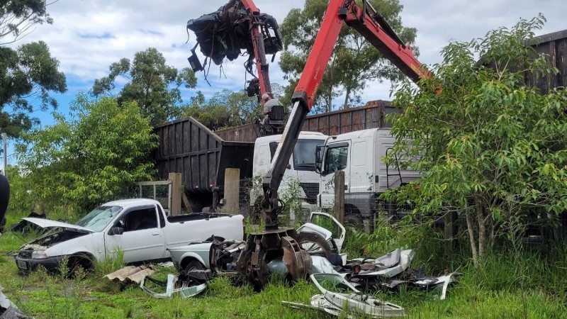 Foto diurna de uma campo com gramado, onde sucatas veiculares estão sendo recolhidas do pátio do estabelecimento e colocadas em caminhões