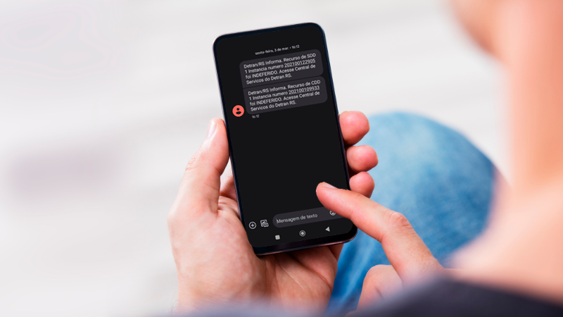 Foto horizontal colorida mostra mãos de uma pessoa branca segurando um celular. Na tela, o exemplo de uma mensagem de texto enviada pelo DetranRS