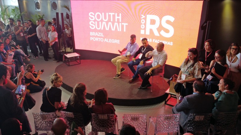 Foto horizontal colorida mostra tres homens sentados em um palco redondo cercado de público. Ao fundo, um painel luminoso traz as marcas do South Summit e do Governo do Estado do RS