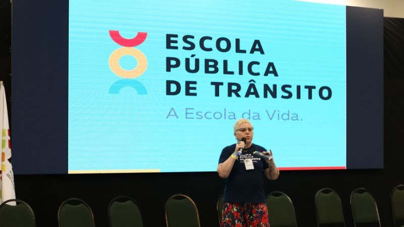 A servidora do DetranRS Carla Badaraco Guglielmi no palco do evento, fazendo sua apresentação. No telão, o logo da Escola Pública de Trânsito da autarquia