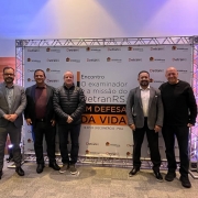 Foto horizontal coloida mostra cinco homens posando para foto em frente a um backdrop com o nome do evento