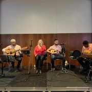 Foto horizontal colorida mostra grupo musical Quarteto Rosa se apresentando em um palco. Mulher loira no meio canta, ladeada por homens com violão, cavaquinho e atabaque