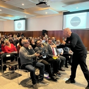 Foto horizontal coloridamostra homem de preto falando ao microfone para um auditório lotado