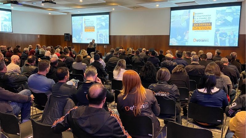 Foto horizontal colorida mostra auditório lotado com pessoas de costas de frente para dois telões