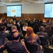 Foto horizontal colorida mostra auditório lotado com pessoas de costas de frente para dois telões
