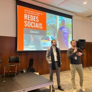 Foto horizontal colorida mostra dois homens se apresentando, um deles falando ao microfone.
