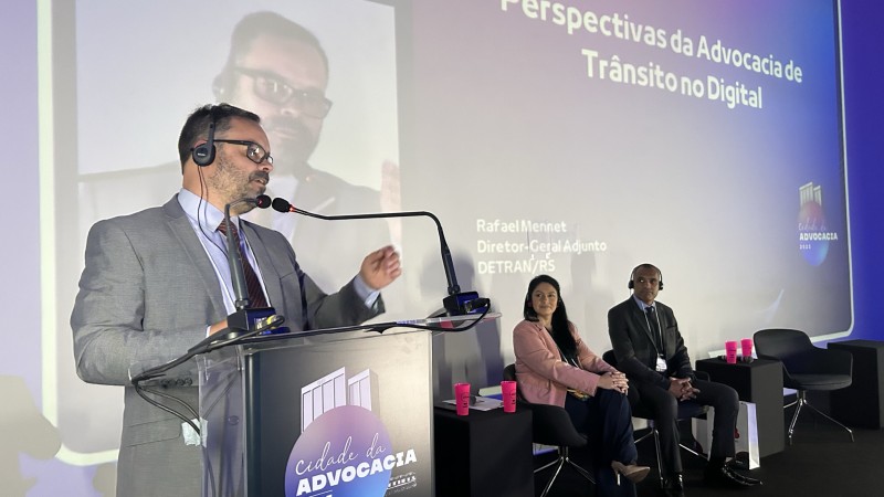 Foto horizontal colorida mostra homem falando em um púlpito. Ao fundo um homem e uma mulher sentados olham para o palestrante