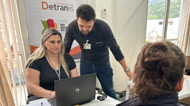 Foto horizontal colorida mostra um homem e uma mulher olhando para a tela de um notebook tendo a frente uma mulher de coque no cabelo. Ao fundo, um banner do DetranRS