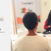 Foto colorida mostra atendente mulher sentada em frente a um notebook falando com um homem que aparece de costas. Ao fundo, um banner com as logos do DetranRS e Governo do RS