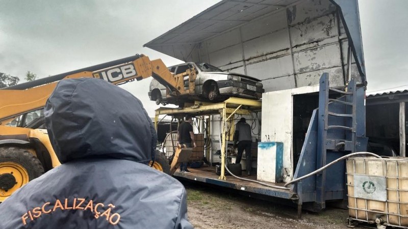 Foto horizontal colorida mostra pessoa de costas com casaco de capuz escrito Fiscalização DetranRS. Homem olha para veículo sendo colocaso por um guindaste em uma plataforma descontaminadora