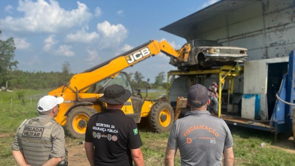 Tres homens uniformizados de costas observam um guindaste elevar uma sucata de carro a uma plataforma móvel de descontaminação