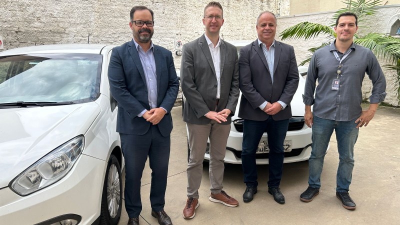Foto horizontal colorida mostra quatro homens posando para foro em frente a dois carros brancos estacionados