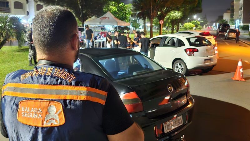 Foto horizontal colorida mostra agente defiscalização da Balada Segura de costas em primeiro plano, com uma blitz de trânsito ao fundo