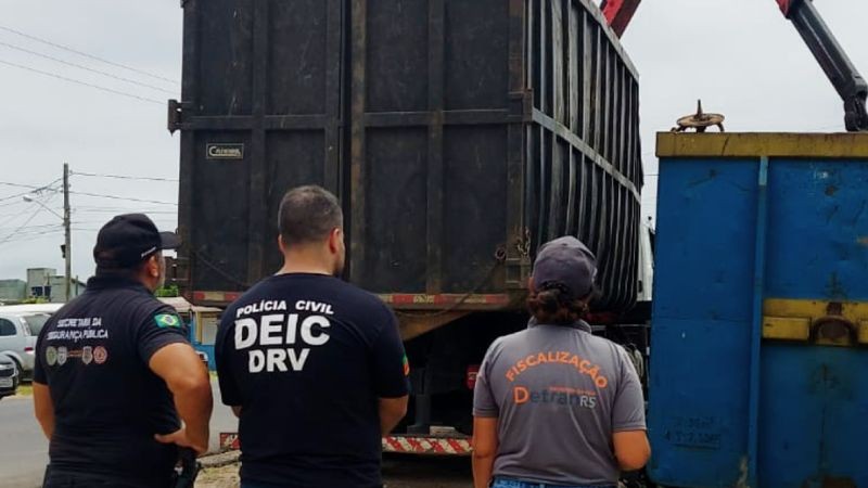 Foto diurna onde servidores da Segurança Pública estão em pé, de costas, olhando máquinas em funcionamento no recolhimento das sucatas apreendidas