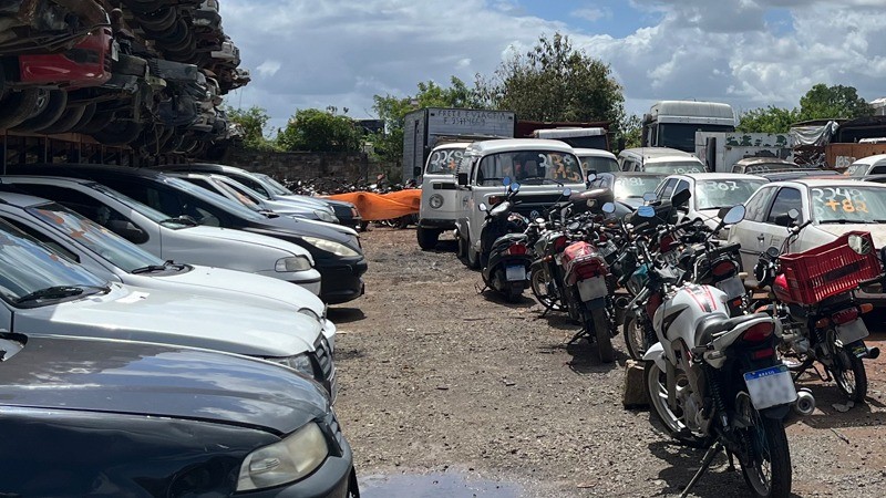 Automóveis usados estacionados lado a lado na esquerda, ao centro motos enfileiradas, ao fundo kombis e à direita mais automóveis, todos para leilão.