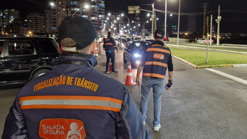 Foto horizontal colorida mostra três agentes de trânsito de costas em um contexto de blitz