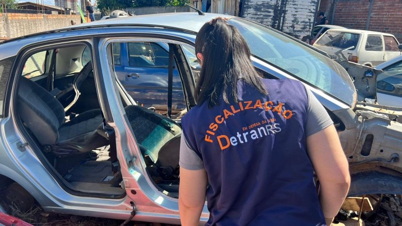 Foto horizontal colorida mostra mulher com colete do DetranRS de costas vistorando uma carcaça de carro durante a operação