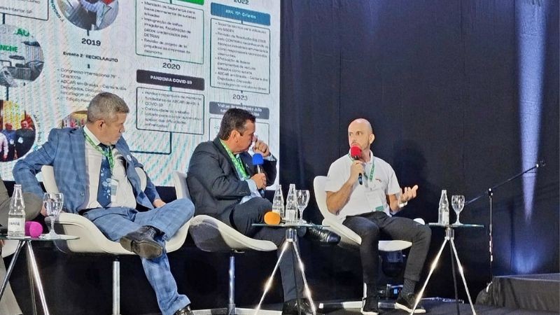 Foto horizontal colorida mostra três homens sentados em cadeiras num palco. O homem da direita fala ao microfone