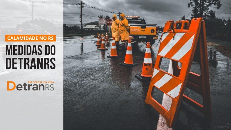 Pista de rodovia interditada por cavalete e cones e agentes de capa de chuva laranja, água em um dos lados da pista.