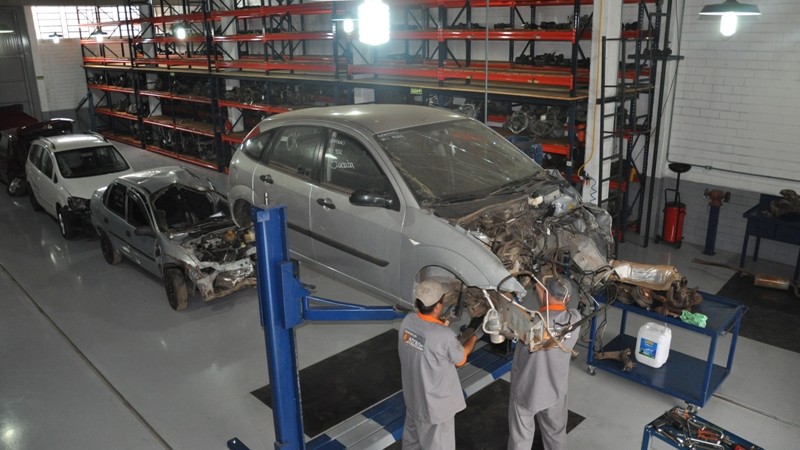 Foto horizontal colorida mostra dois trabalhadores de macacão desmontando um carro elevado por uma plataforma