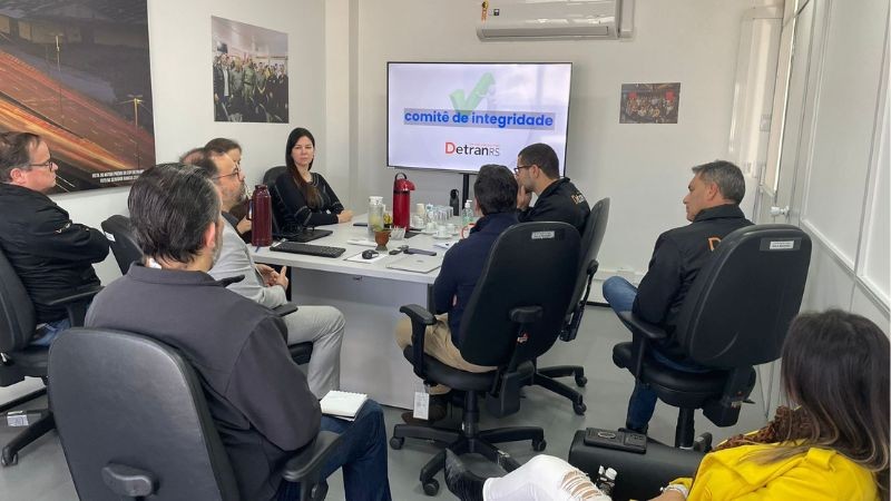 reunião de pessoas em torno de uma mesa retangular e uma tv ao fundo onde está escrito comitê de integridade, detranRS