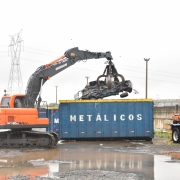 Foto horizontal colorida mostra gancho colocando sucata de veiculo compactada em um container escrito metálicos